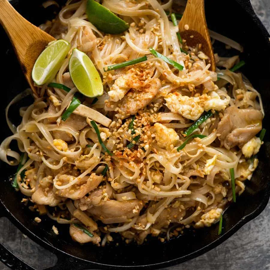 Một đĩa Pad Thai hấp dẫn, với sợi mì gạo vàng óng, tôm, đậu hũ, trứng, cùng các loại rau tươi mơn mởn, phủ lên trên là đậu phộng giã nhỏ và một lát chanh.